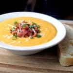 Butternut squash soup topped with bacon, green onion, and parmesan cheese in a white bowl on a wooden board with a piece of bread