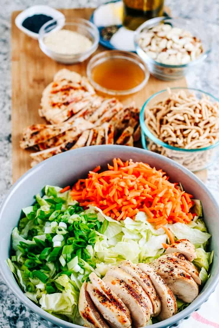 Chinese chicken salad with carrots, green onions and lettuce on a cutting board with dressing, crunchy noodles, almonds and sesame seeds.