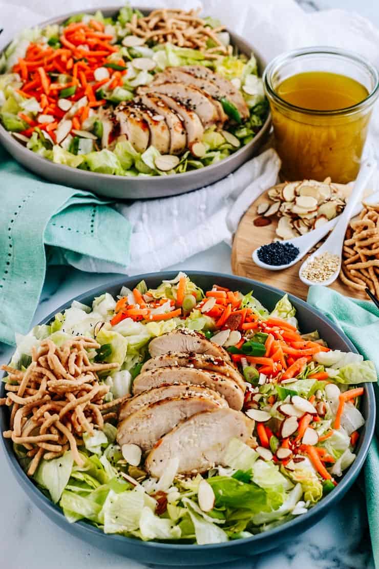 Two Asian salads with grilled chicken in a grey bowls on a white tablecloth with dressing, almonds, and sesame seed next to them. 