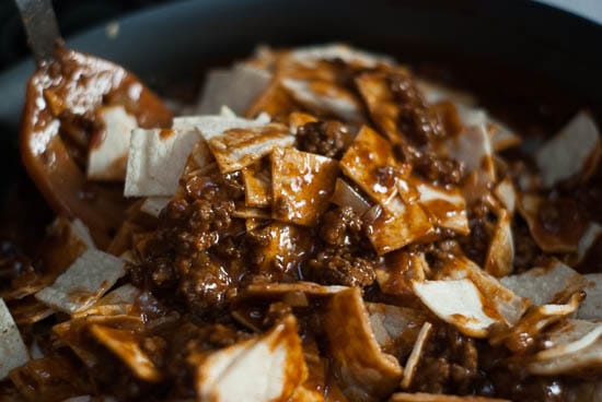 Pouring enchilada sauce and beef over corn tortillas in a skillet.