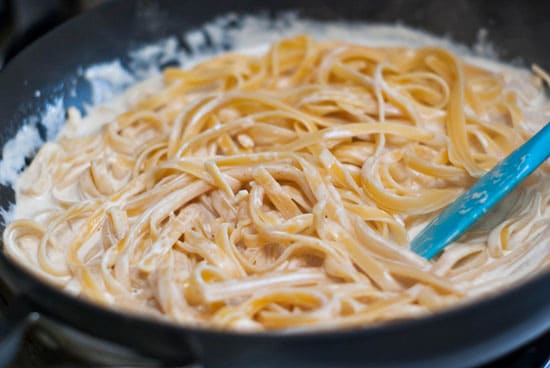 Stirring Brown Butter Fettuccine Alfredo ingredients together in a skillet with a blue spatula.
