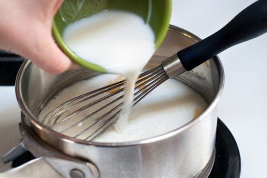 Mixing milk to make this simple Cheese Sauce with a whisk in a silver saucepan.