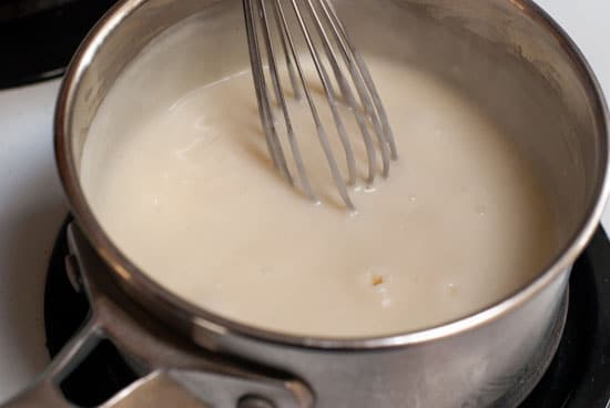 Mixing hot cheese sauce with a silver whisk in a silver saucepan.
