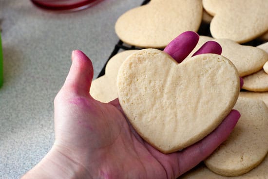 Holding a heart shaped Homemade sugar cookie next to a baking sheet full of warm cookies.