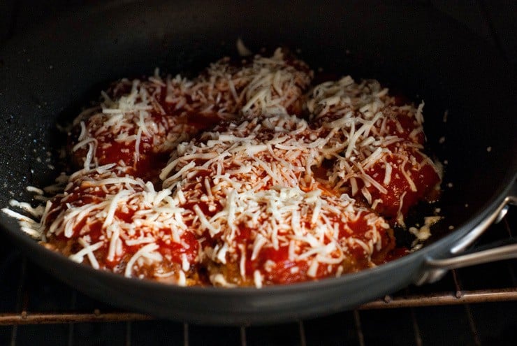 Mozzarella cheese and marinara sauce on beef patties in a black skillet.