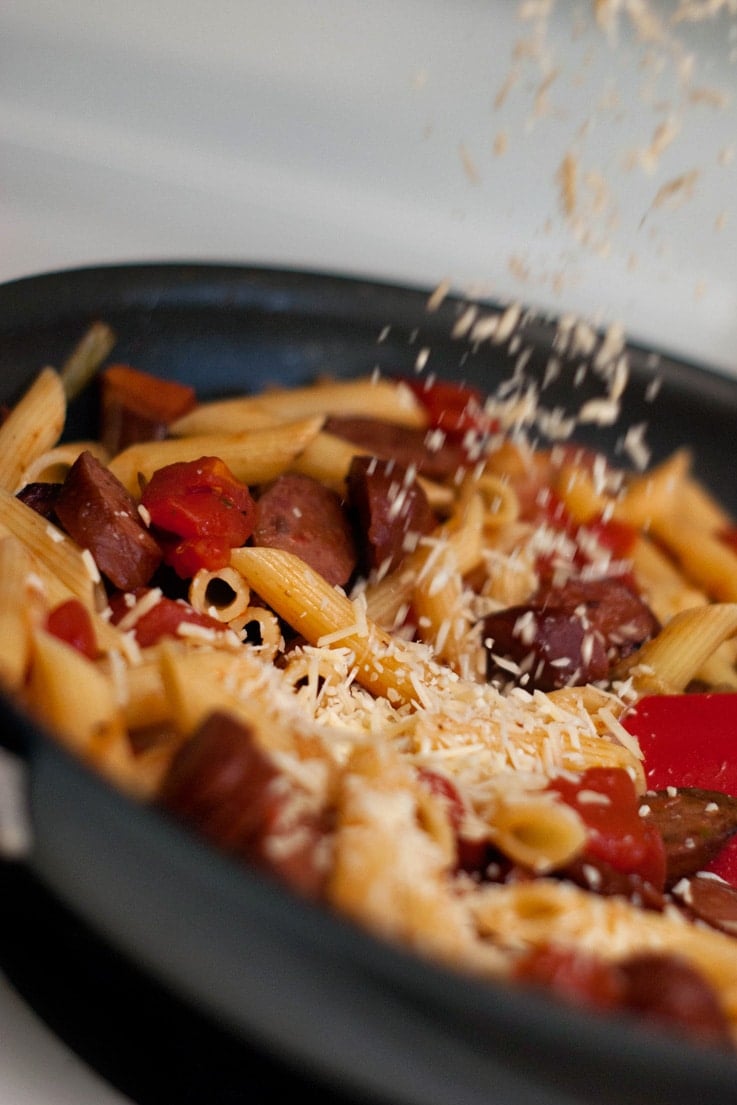 Pouring delicious parmesan cheese onto Penne With Smoked Sausage and Caramelized Onions in a skillet.