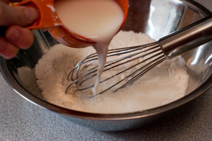 Pouring milk into Boysenberry Cobbler mixture in a silver mixing bowl with a whisk inside.