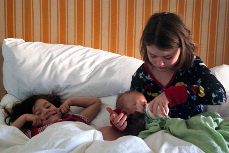 Two young girls feeding bottle to baby in bed.