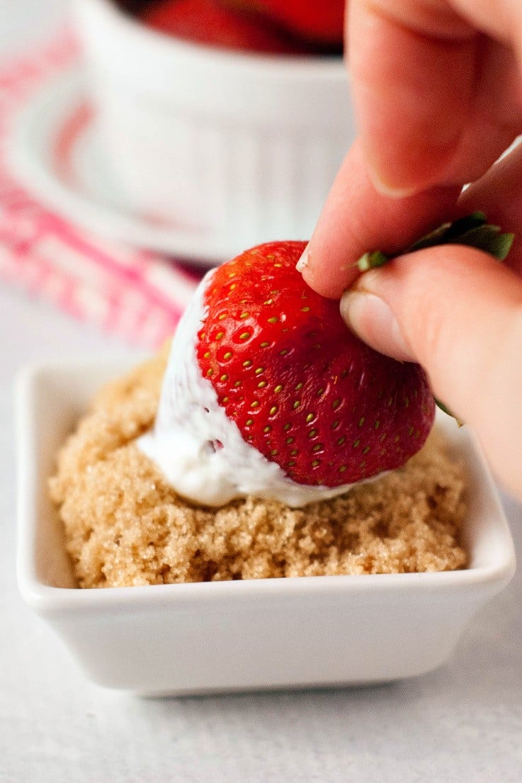 A fresh strawberry being rolled in brown sugar after dipping in sour cream