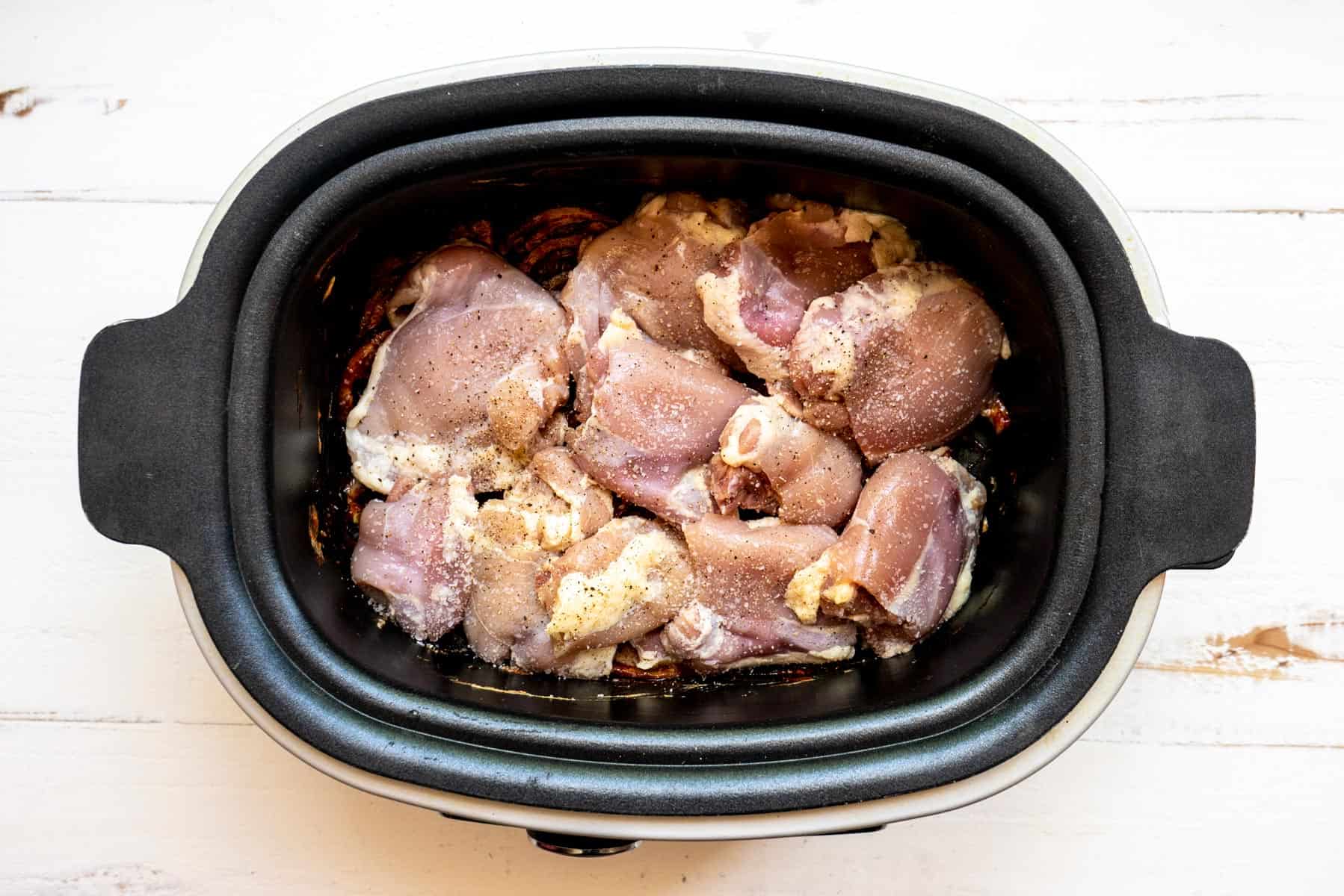 Seasoned chicken thighs in a black slow cooker on a white board