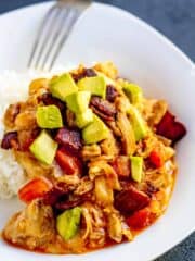 Saucy chicken over rice topped with avocado, bacon and tomato on a white plate with a fork