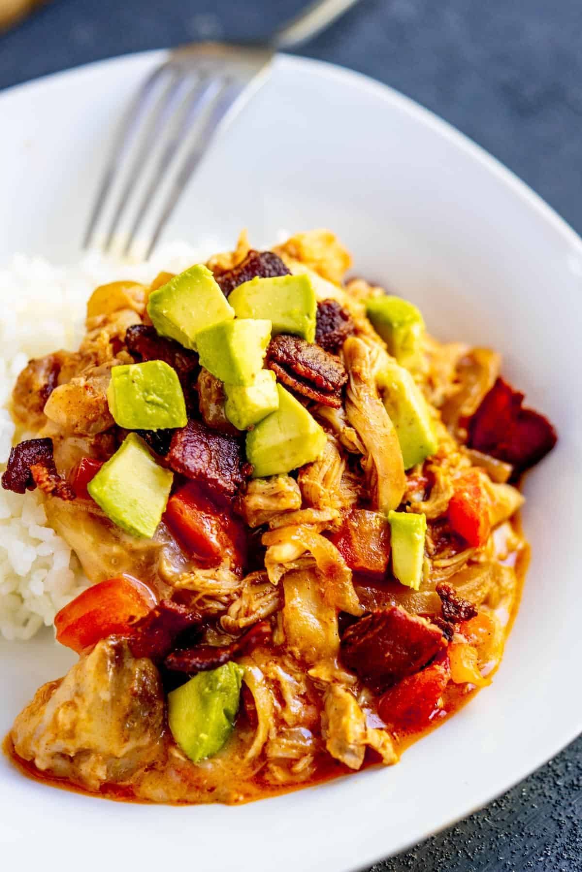 Saucy chicken over rice topped with avocado, bacon and tomato on a white plate with a fork