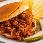 Sloppy Joe on a white plate with pickle and potato chips